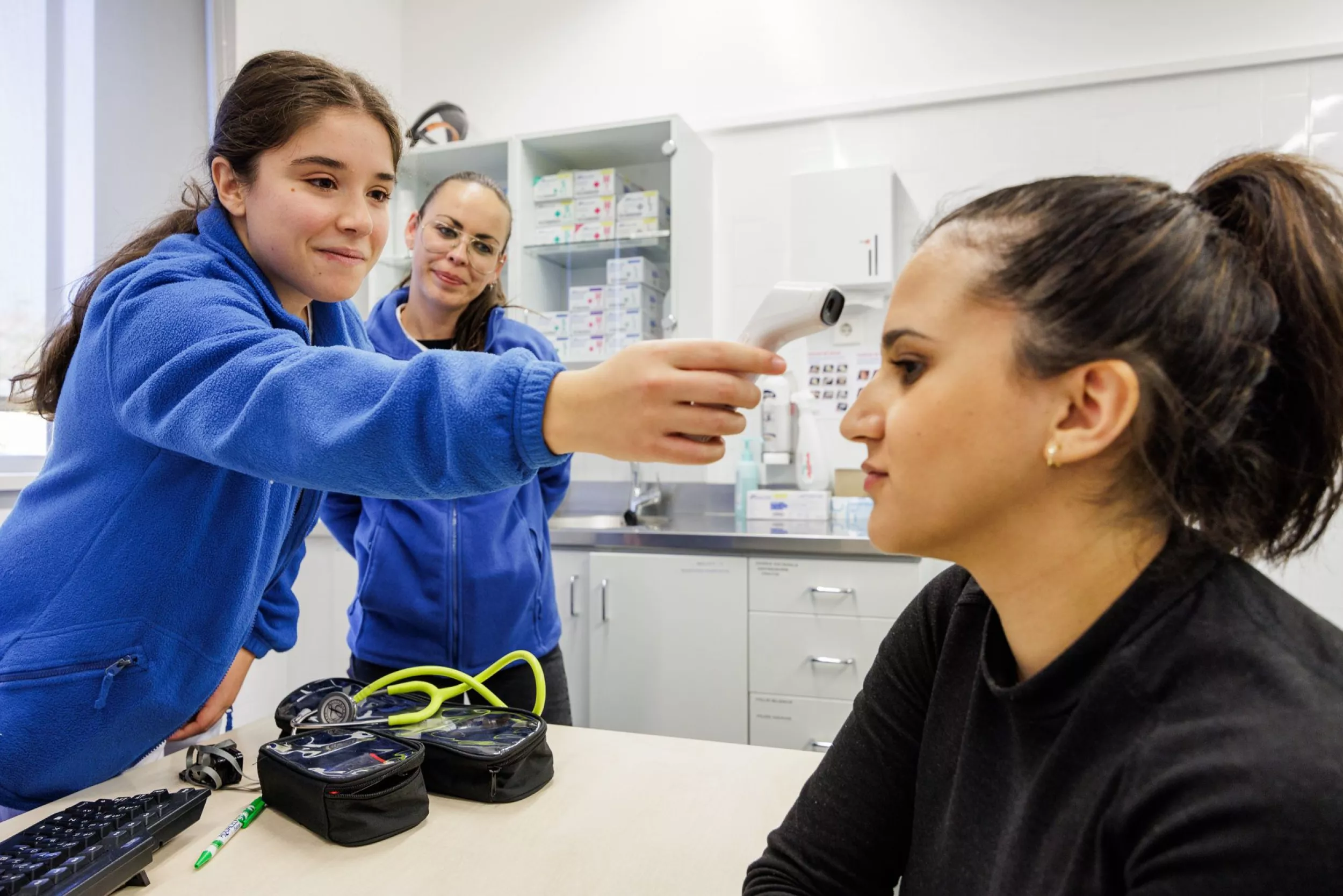 Una enfermera tomándole la fiebre a una paciente, mientras otra enfermera les mira sonriente