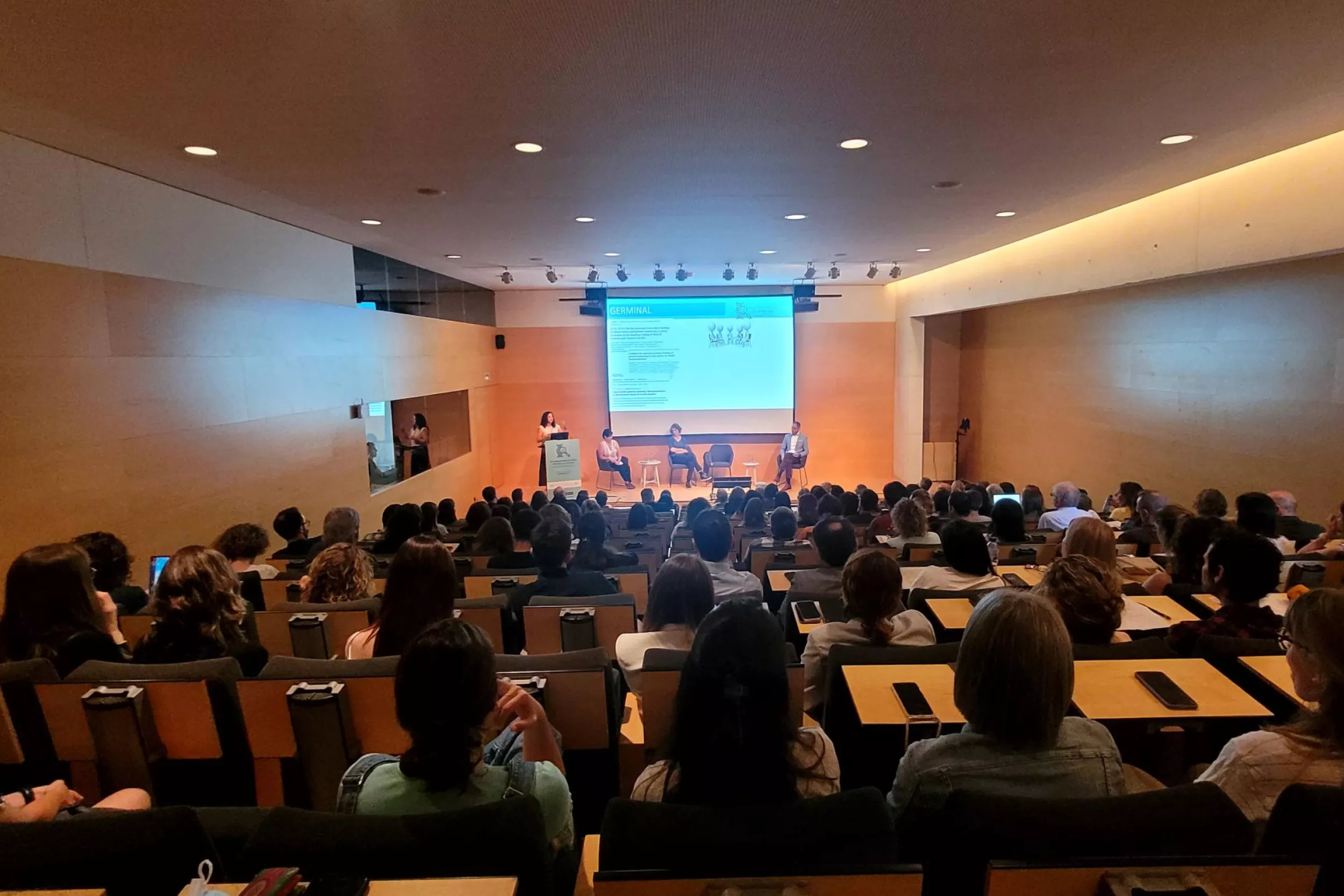 Salón de actos lleno de oyentes, con tres ponentes dando una charla