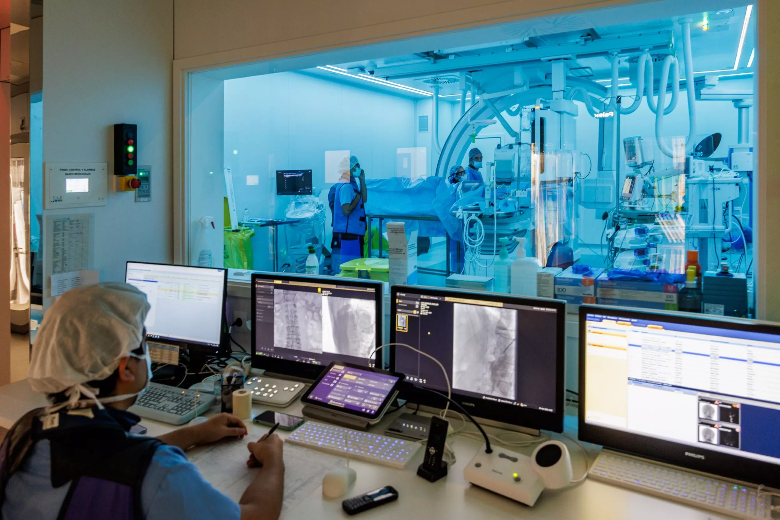 En primer pla es mostra un metge apuntant el que ve a diverses pantalles i al fons es ve una sala de quirofan amb diversos metges.